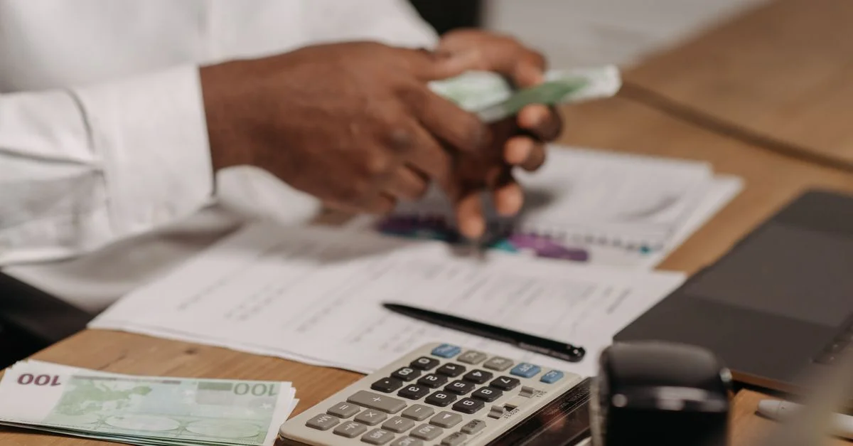 Mains comptant des billets en euros sur un bureau en bois avec une calculatrice, des documents financiers et un ordinateur portable à proximité.