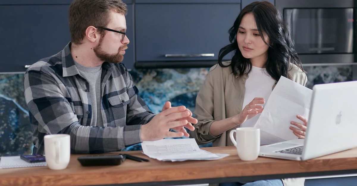 Jeune couple discutant des problèmes financiers avec un ordinateur portable et des documents dans une cuisine moderne.