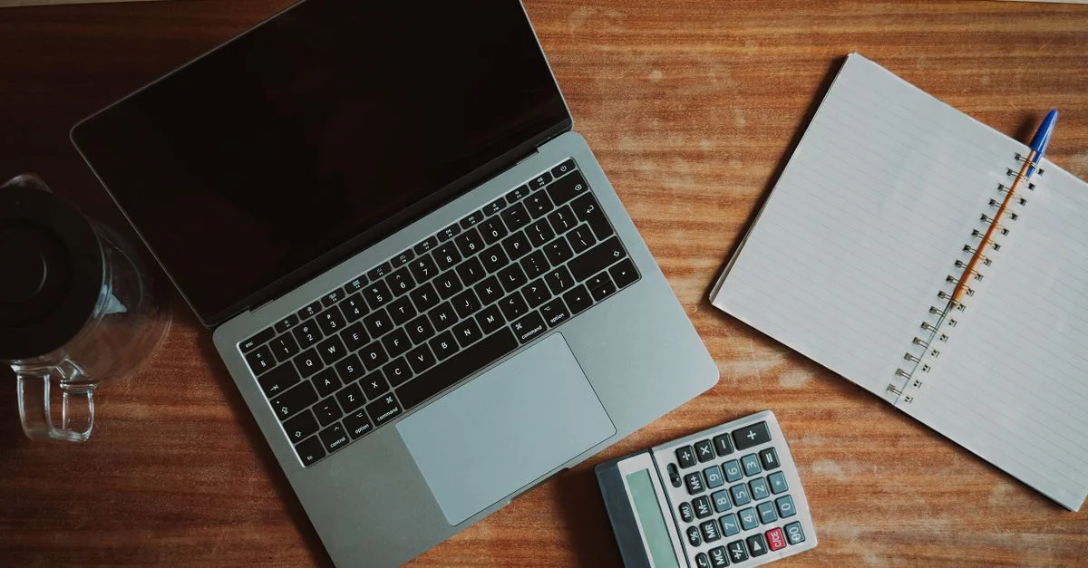 Vue de dessus d'un bureau avec un ordinateur portable, une calculatrice et un bloc-notes ouvert pour le travail ou les études.