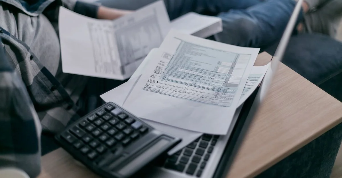 Un jeune adulte utilisant une calculatrice et un ordinateur portable pour gérer ses finances à domicile, entouré de papiers.