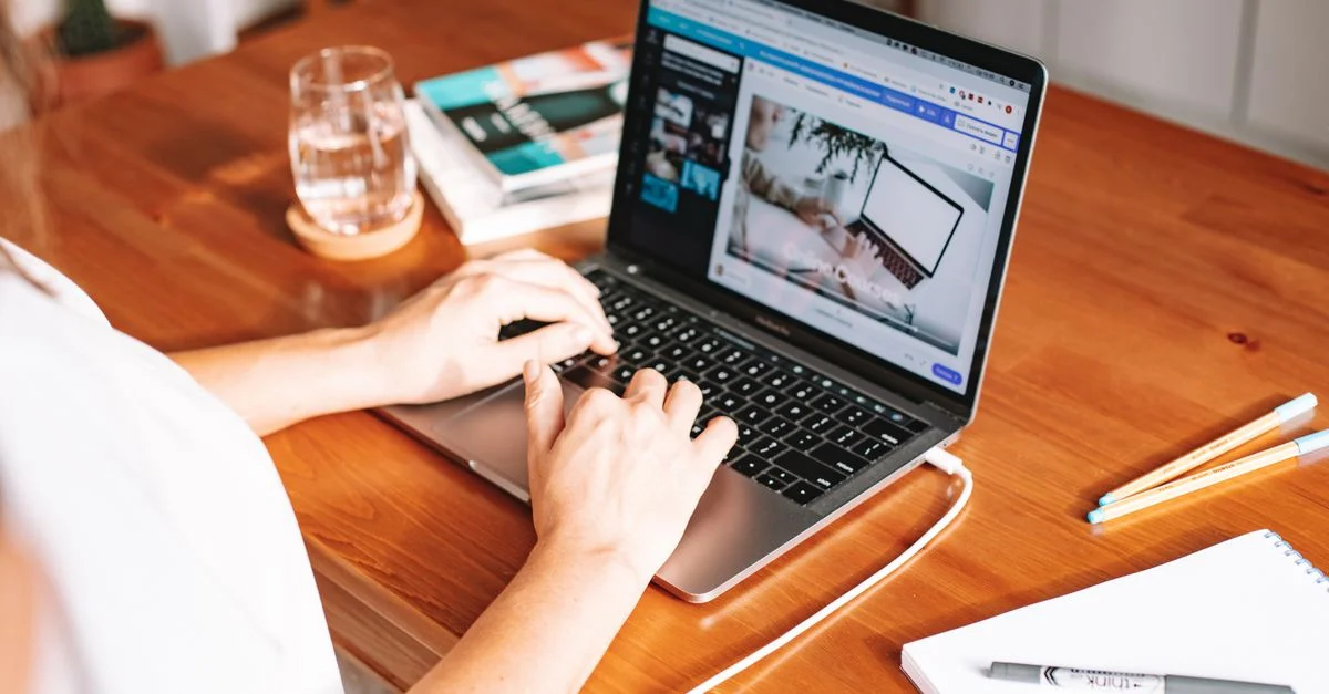 Une personne travaillant à distance sur un ordinateur portable à un bureau en bois avec des fournitures de bureau et un bloc-notes.